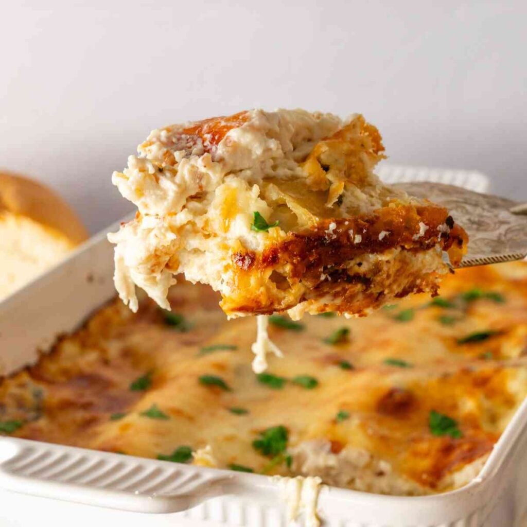 A slice of chicken alfredo lasagna lifted above a pan of hot lasagna.