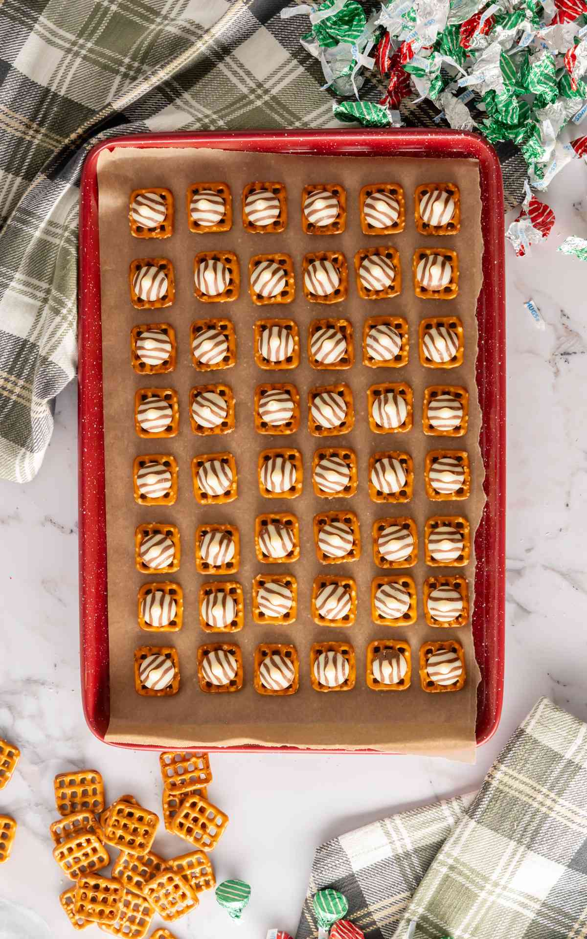 A red baking sheet lined with parchment paper that has square pretzels topped with Hershey hugs.