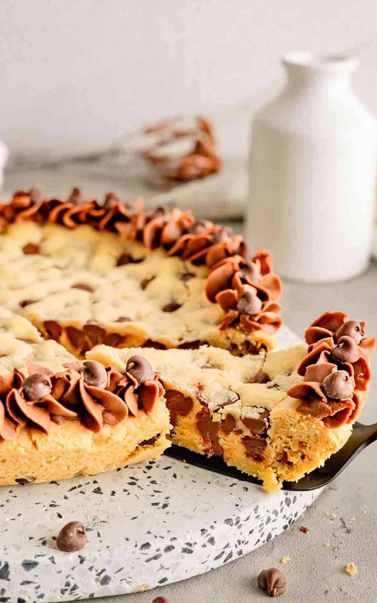 A cake server under a slice of buttercream frosted chocolate chip cookie cake.