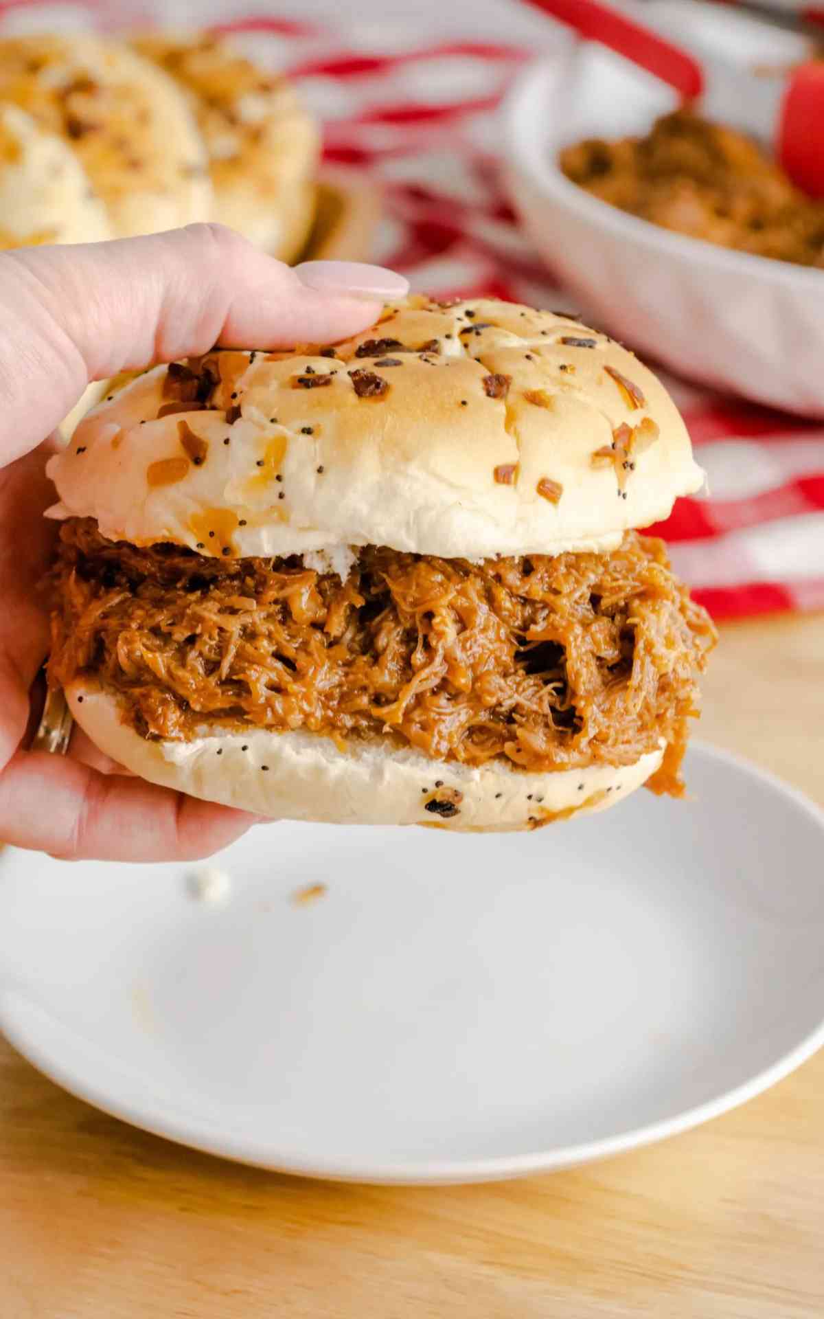 A hold holding up a Root Beer Pulled Pork sandwich on an onion and herb bun with a red checkered linen.