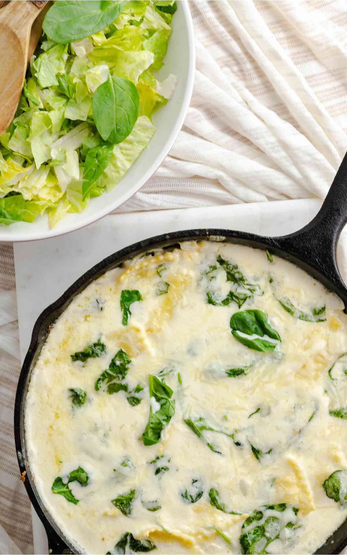 Cast iron skillet with cheese ravioli and spinach with a bowl of salad.