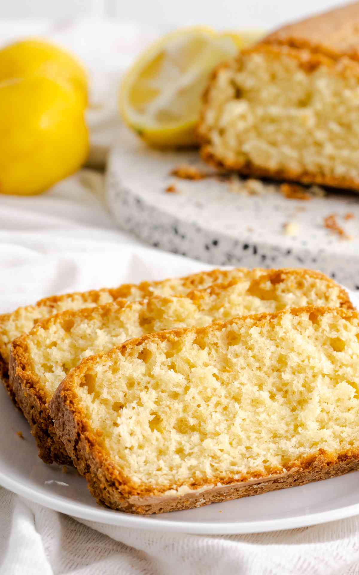 Slices of lemon loaf on a plate with an uncut loaf in the background.