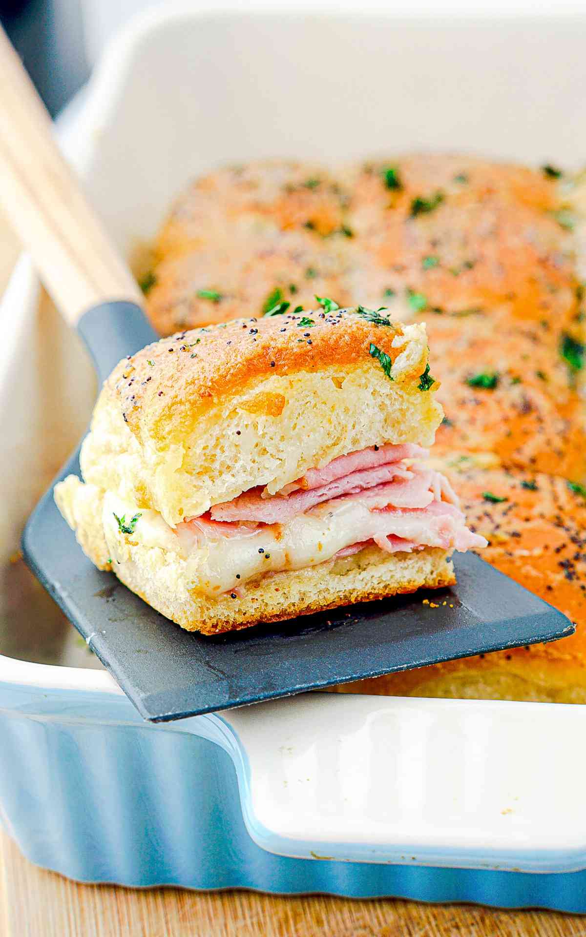 A spatula lifting a ham and cheese slider from a baking dish.
