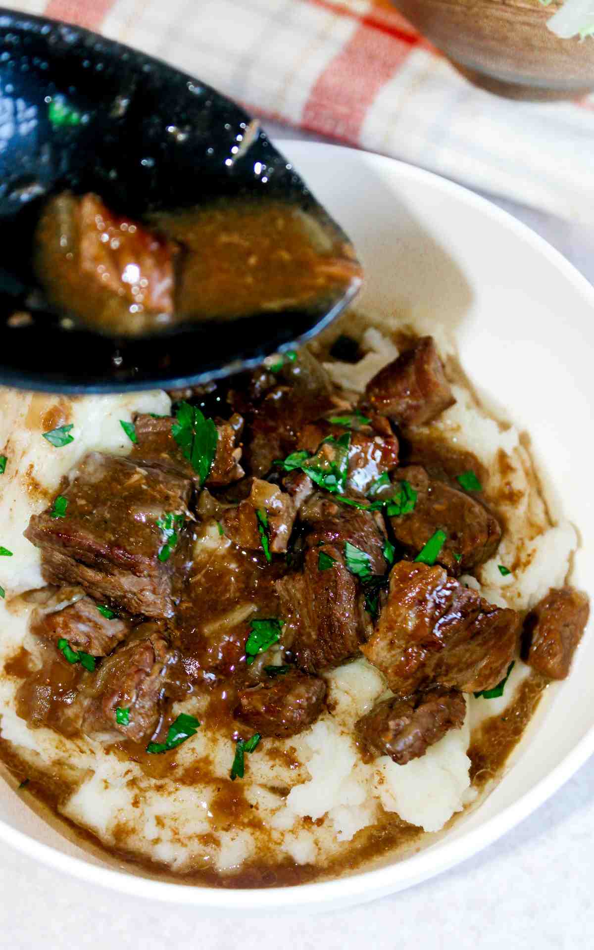 A spoonful of gravy being poured on a bowl of beef tips and mashed potatoes.